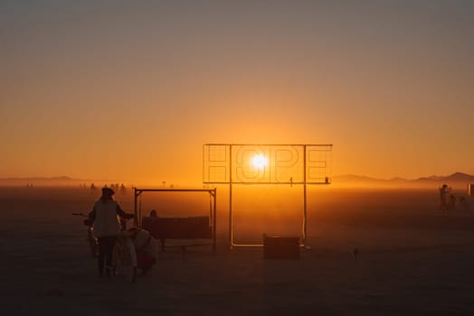 The sun sets behind a large HOPE installation at a desert festival, aligning with the letter O. Silhouettes of participants and distant mountains are visible.