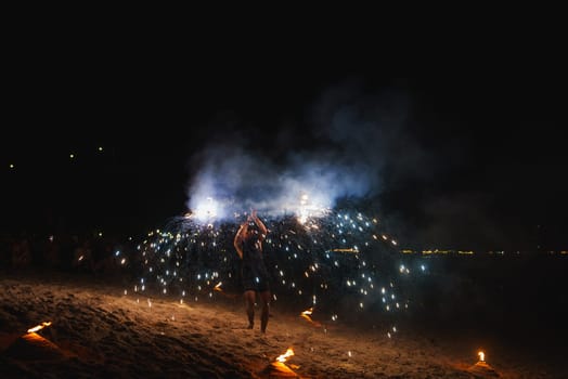 A performer spins fire on a sandy beach at night, with torches in the foreground and faint lights from distant structures or boats in the background.