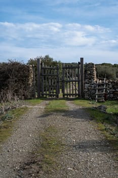 A rustic wooden gate with dry stone walls, located in a rural countryside with a dirt path and natural vegetation. The setting conveys tranquility and authenticity