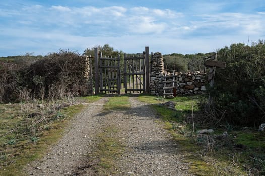 A rustic wooden gate with dry stone walls, located in a rural countryside with a dirt path and natural vegetation. The setting conveys tranquility and authenticity