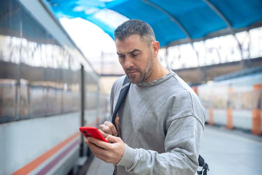 Serious man wearing a gray sweatshirt holding a red smartphone while looking around at a train station. His expression conveys anticipation, travel, and modern connectivity.