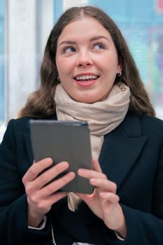 Focused young woman in a black coat and beige scarf sitting inside a modern subway train, reading on an e-reader. She enjoys a quiet moment during her daily commute.