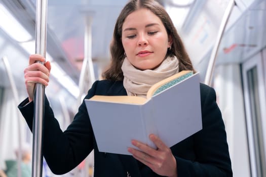 Focused young woman in a black coat and beige scarf standing inside a modern subway train, reading a book while holding onto a metal pole. She enjoys a quiet moment during her commute.