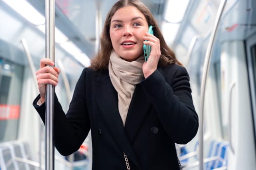 Cheerful young woman in a black coat and beige scarf talking on her smartphone while standing inside a modern subway train. She smiles brightly, holding onto a metal pole during her commute.