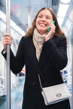 Cheerful young woman in a black coat and beige scarf talking on her smartphone while standing inside a modern subway train. She smiles brightly, holding onto a metal pole during her commute.