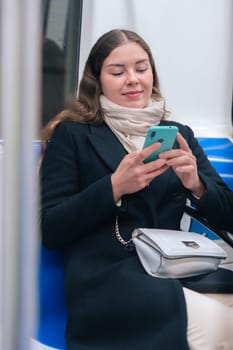 Cheerful young woman in a black coat and beige scarf sitting inside a modern subway train, laughing while looking at her smartphone. She enjoys a fun moment during her commute.