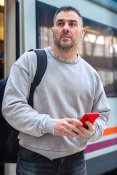 Serious man wearing a gray sweatshirt holding a red smartphone while looking around at a train station. His expression conveys anticipation, travel, and modern connectivity.