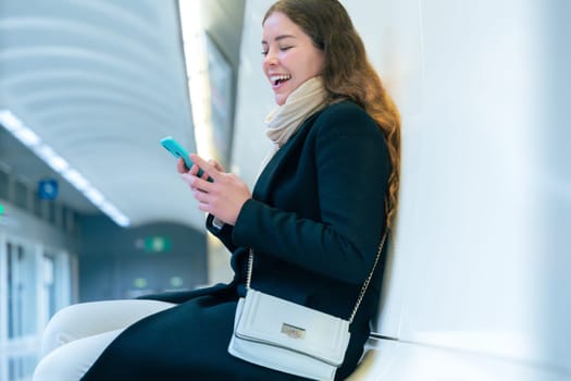 Stylish young woman in a black coat and scarf sitting in a modern subway station, smiling while using her smartphone. The bright and clean environment enhances the urban setting.
