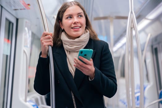 Young woman in a black coat and beige scarf standing inside a modern subway train, holding a smartphone and gripping a metal pole. She appears focused while commuting, carrying a stylish white purse.