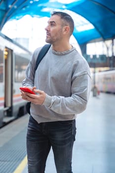 Serious man wearing a gray sweatshirt holding a red smartphone while looking around at a train station. His expression conveys anticipation, travel, and modern connectivity.