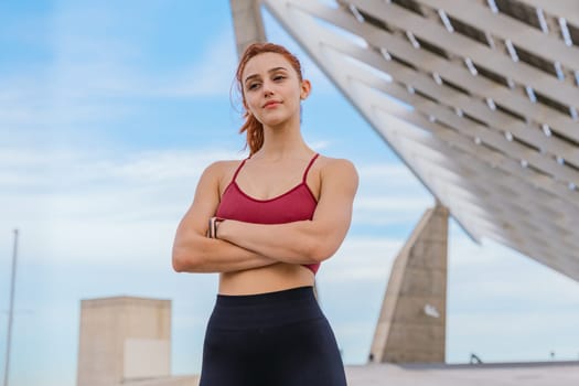 Young fit woman with red hair wearing sportswear, standing confidently with arms crossed in an urban setting. Modern architectural structure in the background under a blue sky.