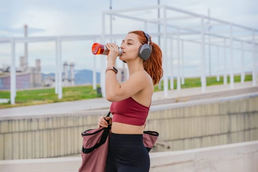 Athletic red-haired woman wearing sportswear and headphones, drinking water after exercise. She carries a gym bag and stands in a modern outdoor urban environment under a cloudy sky.