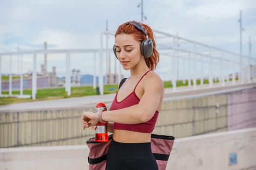 Fit young woman with red hair wearing sportswear and headphones, checking her smartwatch while holding a water bottle. She carries a gym bag and stands in a modern outdoor urban environment.