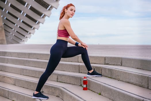 Athletic red-haired woman wearing sportswear stretching her leg on outdoor stairs in a modern urban environment. A water bottle sits nearby as she prepares for exercise under a bright blue sky.