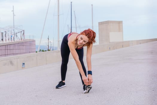 Fit red-haired woman wearing sportswear bending forward to stretch her leg in an outdoor urban setting. She prepares for exercise under a cloudy sky, maintaining flexibility and strength.