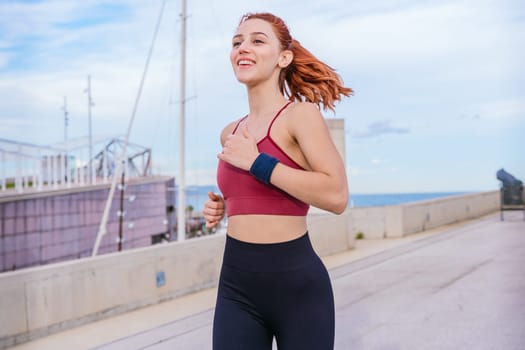 Fit young woman with red hair running outdoors near the coast. She is wearing sportswear and looks happy and energetic. The background features a bright sky and a modern urban environment.