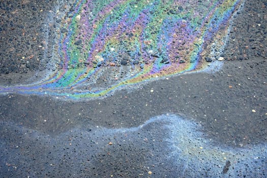 An iridescent oil spill on the asphalt road showcases colorful designs, indicating both environmental contamination and a risk for motorists.