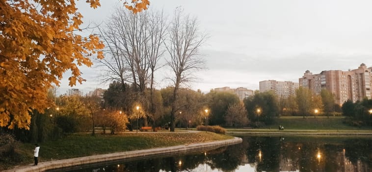 A tranquil city park features a reflective lake surrounded by autumn trees and city buildings. Soft evening light illuminates the landscape as the day transitions into night.