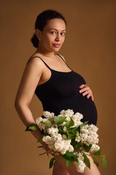 A pregnant woman stands gracefully holding a bouquet of white flowers, exuding beauty and the essence of motherhood, against a soft brown background. The scene captures anticipation, serenity, and love.