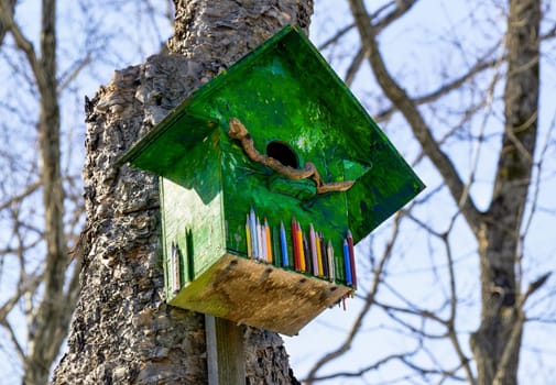 Green wooden birdhouse with 
color pencils. It hanging on the tree in forest on sky background. Early spring, Russia