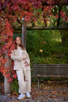 A young woman with wavy light brown hair and a cheerful smile is leaning against a wooden pillar wrapped in red autumn leaves. She is dressed in a beige casual outfit, including a stylish denim jacket and matching pants, with a white top underneath. The background features a scenic garden pergola with wooden beams covered in lush green and red foliage, creating a natural and cozy atmosphere. The warm lighting and soft depth of field enhance the autumn aesthetic, making this image perfect for seasonal fashion, lifestyle blogs, outdoor portraiture, and autumn-themed marketing campaigns. Smiling young woman in beige casual outfit leaning on wooden pillar with red autumn leaves in scenic garden pergola – warm and cozy fall portrait with natural background