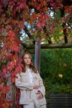 Capture the essence of autumn with this charming portrait of a young woman embracing the season's warmth and beauty. The subject, dressed in a casual beige outfit, leans comfortably against a wooden pillar of a garden pergola, her smile radiating contentment and joy. Vibrant red autumn leaves frame the scene, creating a stunning natural backdrop that epitomizes fall's rich palette. The rustic pergola adds a touch of countryside charm, while the overall composition exudes a cozy, inviting atmosphere. This image is perfect for autumn-themed marketing campaigns, lifestyle blogs, fashion editorials, and travel publications, showcasing the beauty of outdoor living and seasonal transitions.