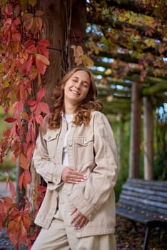A young woman with wavy light brown hair and a cheerful smile is leaning against a wooden pillar wrapped in red autumn leaves. She is dressed in a beige casual outfit, including a stylish denim jacket and matching pants, with a white top underneath. The background features a scenic garden pergola with wooden beams covered in lush green and red foliage, creating a natural and cozy atmosphere. The warm lighting and soft depth of field enhance the autumn aesthetic, making this image perfect for seasonal fashion, lifestyle blogs, outdoor portraiture, and autumn-themed marketing campaigns. Smiling young woman in beige casual outfit leaning on wooden pillar with red autumn leaves in scenic garden pergola – warm and cozy fall portrait with natural background