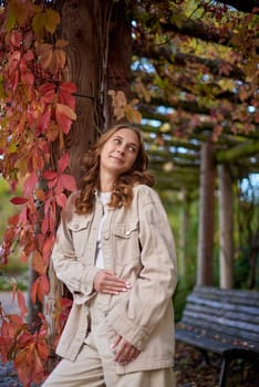 A young woman with wavy light brown hair and a cheerful smile is leaning against a wooden pillar wrapped in red autumn leaves. She is dressed in a beige casual outfit, including a stylish denim jacket and matching pants, with a white top underneath. The background features a scenic garden pergola with wooden beams covered in lush green and red foliage, creating a natural and cozy atmosphere. The warm lighting and soft depth of field enhance the autumn aesthetic, making this image perfect for seasonal fashion, lifestyle blogs, outdoor portraiture, and autumn-themed marketing campaigns. Smiling young woman in beige casual outfit leaning on wooden pillar with red autumn leaves in scenic garden pergola – warm and cozy fall portrait with natural background