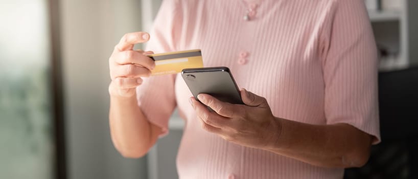 An elderly woman using her smartphone to shop online, holding a yellow credit card in one hand, showcasing a modern shopping experience in her cozy home.
