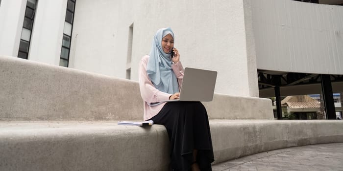 A Muslim business woman wearing a hijab engages in a phone conversation while working on her laptop in an outdoor setting.