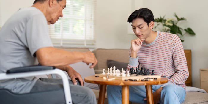 A son deeply focused on his next move in a chess game with his father, who is in a wheelchair, showcasing their close family bond during leisure time together.