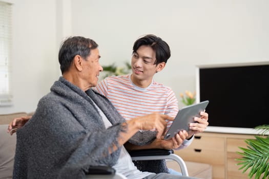 A caring son interacts with his elderly father, who is wrapped in a blanket, as they explore a tablet together, fostering connection and joy in their home.