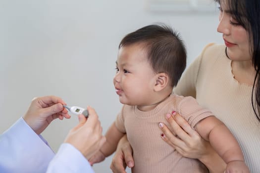 A curious baby reacts while a pediatrician measures their temperature with a thermometer, as the mother supports them in a bright medical clinic, ensuring health.