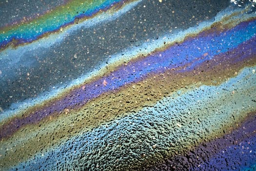An overhead view of an asphalt road showing a large, iridescent slick of petrol creating a large polluted area