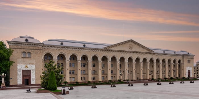Panoramic view of Ganja City Hall, in front of spacious plaza, situated in Azerbaijan, against backdrop of sunset, characterized by its beige color, arched facade, and numerous windows