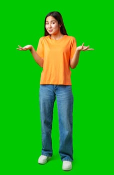 A young woman wearing an orange t-shirt and blue jeans stands with her hands raised, displaying a playful, unsure expression. The bright green background adds vibrancy to the atmosphere.