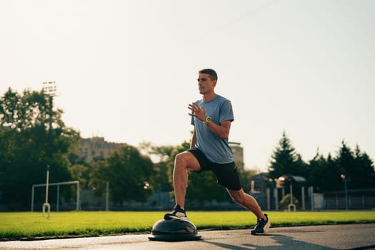 A man engages in lunges on a balance trainer as part of his workout routine at a park. The sun sets in the background, creating a vibrant evening atmosphere for fitness.