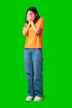 A young woman stands in a bright green environment, covering her face with her hands in an expression of surprise or shyness. She wears an orange shirt and blue jeans.