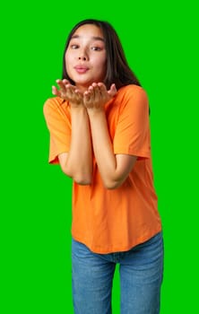 A young woman is playfully blowing kisses while wearing an orange t-shirt and blue jeans against a vibrant green background, capturing a fun and lively moment.