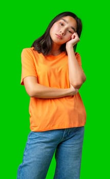 A young woman is standing in a relaxed pose, wearing an orange t-shirt and blue jeans. She rests her chin on her hand, displaying a thoughtful expression, within a vibrant green studio backdrop.