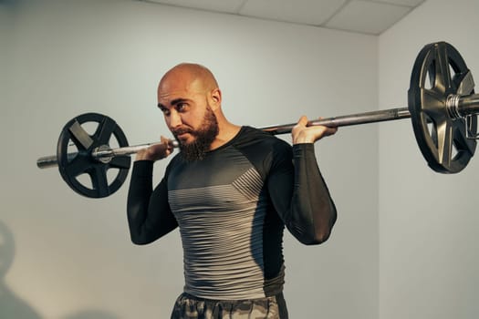 A man with a beard and bald head performs a barbell squat in a gym, showcasing his strength and focus. The workout occurs in a well-lit space early in the day.