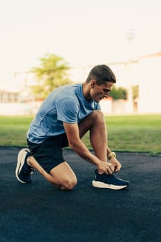 A young man kneels on a track field, tying his running shoes as he prepares for a workout. The soft evening light casts a warm glow over the scene, enhancing his focus and determination.