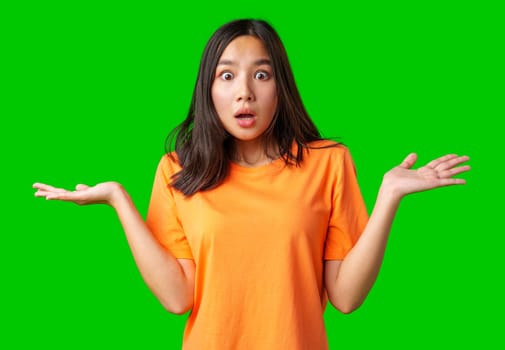 A young woman is standing with a surprised expression, wearing an orange shirt. Her hands are raised in a questioning manner against a vivid green background.