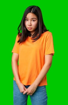 A young woman stands in an orange shirt and blue jeans with her hands clasped in front. Her long hair frames her face as she poses against a vivid green backdrop.
