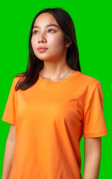 A young woman stands confidently with a slight gaze upwards, wearing a vibrant orange t-shirt. The bright green background enhances the vivid colors, creating a dynamic setting.