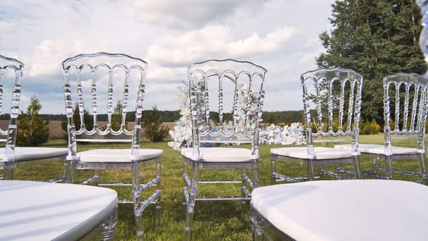 Wedding setting featuring transparent chairs, white cushions scattered across green lawn beneath overcast sky, preparing for ceremonial moment