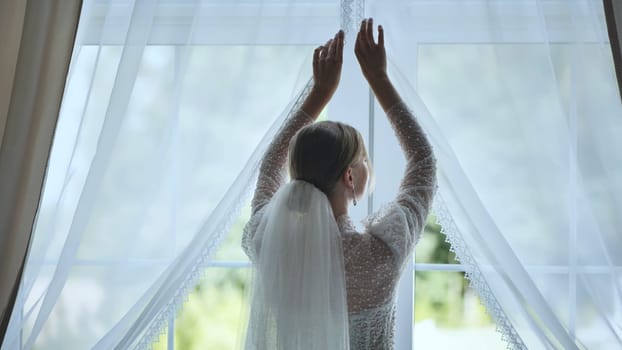 Bride in flowing wedding dress revealing sunlit window, soft veil trailing, capturing serene morning wedding preparation moment