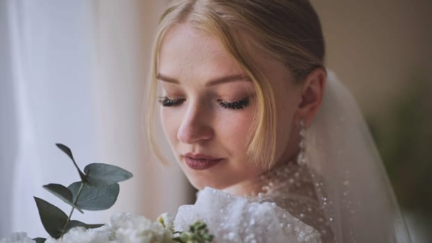 Elegant bride standing near window, holding white flower bouquet, soft natural lighting illuminating bridal attire