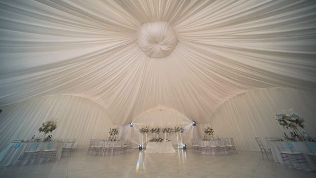 Elegant tent decorated with white ceiling drapery and flower arrangements, set up for a wedding reception, creating a romantic and luxurious atmosphere