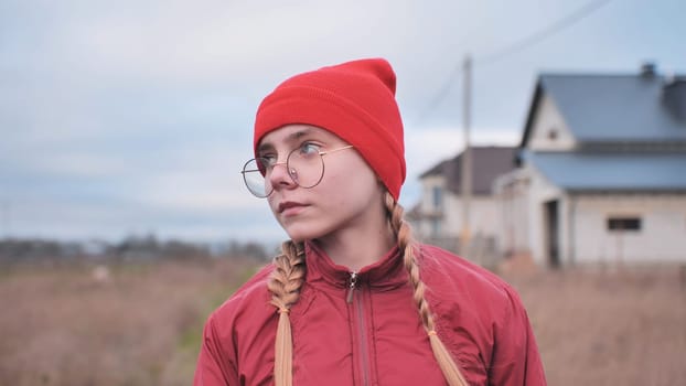 Teenage girl wearing red beanie and glasses, standing contemplatively in grassy field, gazing into distance